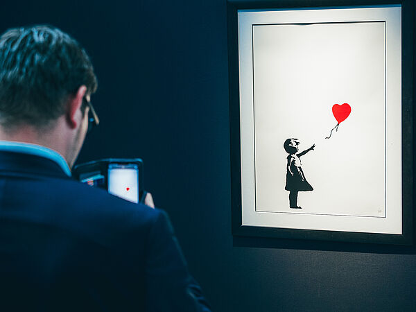 Ein Mann steht vor einem Bild. Auf dem Bild ist ein Mädchen zu sehen, das einen Ballon in der Hand hält.