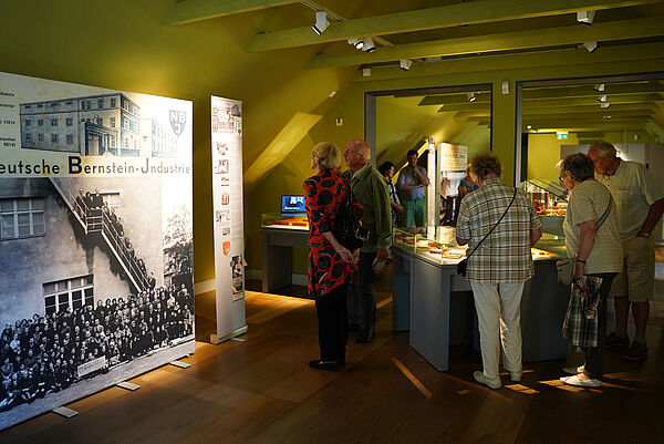 Menschen stehen um eine Vitrine. Links ein Plakat zur Ausstellung.