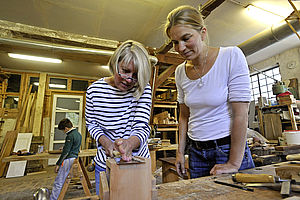 Zwei Frauen stehen in einer Werkstatt nebeneinander an einer Werkbank. Eine von ihnen 