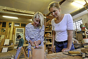 Zwei Frauen stehen in einer Werkstatt nebeneinander an einer Werkbank. Eine von ihnen 