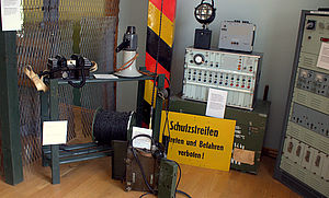 Ein Funkgerät, eine tragbare Lampe, ein Megafon, ein gelbes Schild mit der Aufschrift "Schutzstreifen. Betreten und Befahren verboten!", ein Grenzpfahl und mehrere technische Gegenstände. 