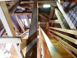 Eine Treppe. Rundherum Holzgebälk. Die Perspektive blickt von oben nach unten.