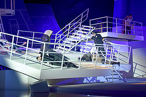 Das Deck kippt nach vorn. Schauspieler liegen auf der Bühne oder stemmen sich am Geländer gegen die Bewegung. 