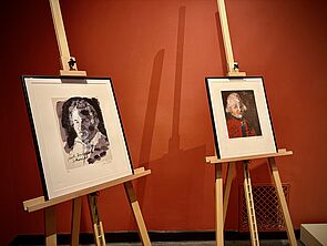 Zwei Bilder stehen auf Staffeleien vor der roten Wand.