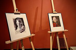 Zwei Bilder stehen auf Staffeleien vor der roten Wand.