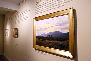 Ein Landschaftsbild im goldenen Rahmen hängt an einer Wand.