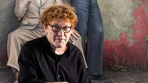 Manfred W. Jürgens sitzt vor einem seiner Gemälde. Er hält zwei Pinsel und eine Mischpalette in der Hand. 