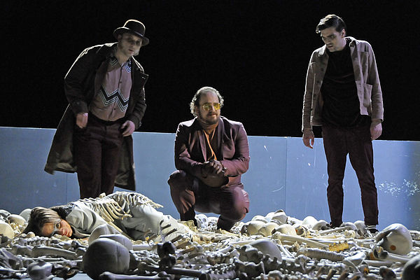 Brunhild liegt auf der Bühne zwischen Knochen und Skeletten. Drei Schauspieler stehen vor ihr und betrachten sie.
