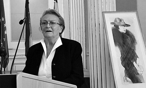 Eine Frau steht an einem Rednerpult. Im Hintergrund steht ein Bild auf einer Staffelei.