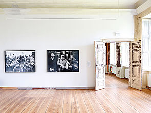 Ein heller Raum. Rechts befindet sich eine geöffnete Flügeltür, deren Farbe hier und da schon abgeplatzt ist. An der Wand hängen zwei Schwarz-Weiß-Fotografien.