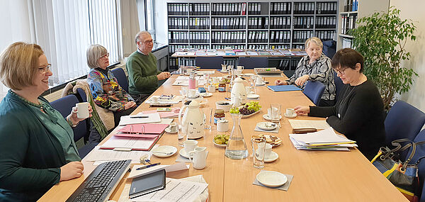 An einem Konferenztisch sitzen vier Frauen und ein Mann. Sie unterhalten sich. Vor ihnen liegen Mappen und lose Blätter. Auf dem Tisch stehen Tassen und Kaffeekannen.