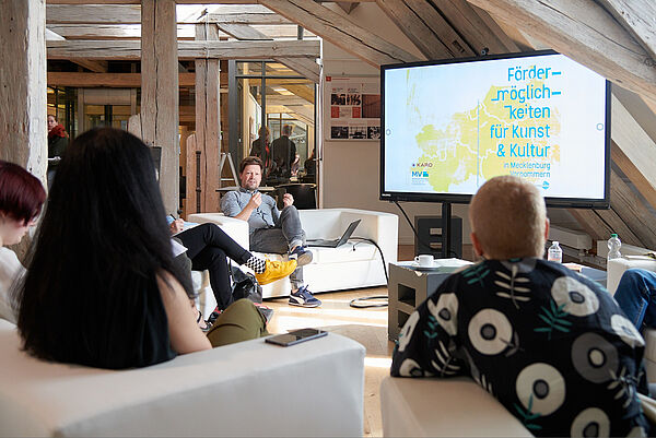 Ein Raum mit Dachschrägen und Balken. Drei Jugendliche sitzen mit dem Rücken zum Foto auf Sesseln. Sie schauen zu Martin Auer. Er sitzt auf einem Sofa und spricht. Im Raum steht ein großer Bildschirm. Darauf steht "Fördermöglichkeiten für Kunst und Kultur".