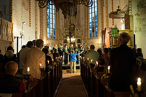 Blick in einer Kirche von hinten auf die Bühne. Rechts und links sind Gäste zu sehen.