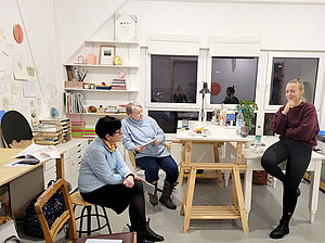 Zwei Frauen sitzen an einem Tisch in einem Atelier. Die Künstlerin sitzt auf dem Tisch und erzählt. 