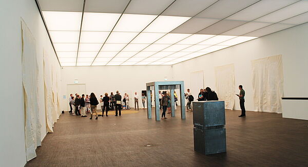 Ein Ausstellungsraum. An den Wänden hängen weiße Tücher. Im Raum stehen ein XXL-Tisch und ein Sockel. Frauen und Männer gehen durch den Raum.