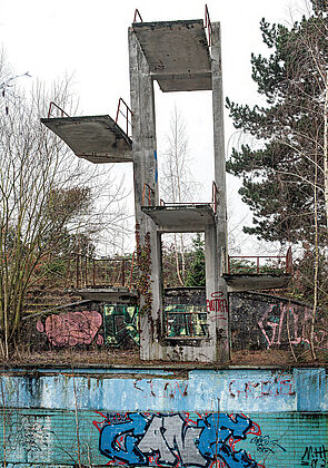 Ein verfallener Sprungturm am Rande eines verwitterten Freibandbeckens. 