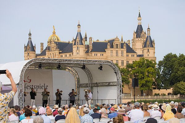 Ein Publikum sitzt vor einer Bühne mit Musikern. Im Hintergrund steht das Schweriner Schloss.