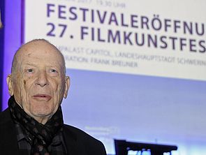 Wolfgang Kohlhaase steht in einem der Kinos vor einer Leinwand. 