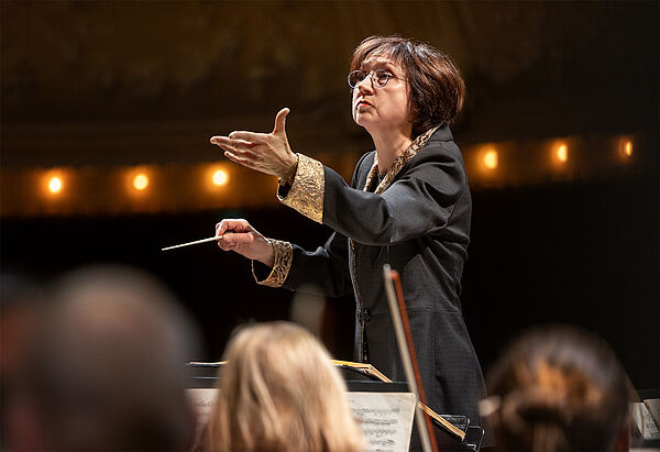 Eine Frau steht vor einem Orchester und dirigiert. 