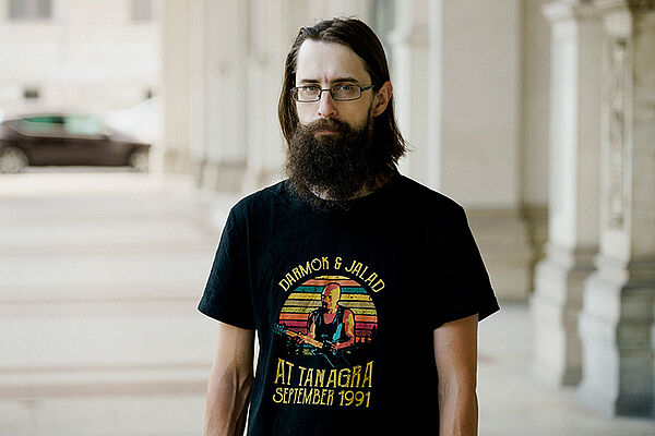 Ein Mann mit Vollbart und Brille. Er steht vor einem Gebäude. Das Gebäude ist verschwommen.