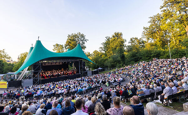Eine Bühne. Die Bühne steht in einem Park. Auf der Bühne sind Musiker. Vor der Bühne sitzen viele Menschen.