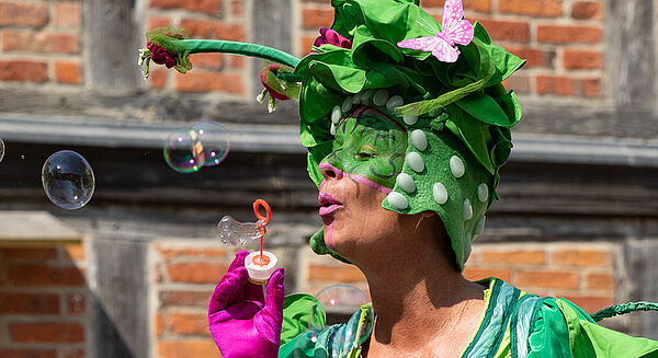 Ein maskiertes Gesicht. Die Person trägt einen Hut wie eine Blume. Sie macht Seifenblasen.