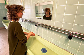 An der Wand reiht sich Waschbecken an Waschbecken. An den Wasserrohren sitzen in regelmäßigen Abständen Wasserhähne. An einem von ihnen wäscht Lisa Marie Steude ihre Hände und schaut in den Spiegel.