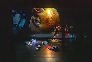Fünf Schauspieler liegen auf dem Boden. Auf der Wand ist ein großer Kopf auf einer Leinwand zu sehen.