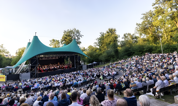 Zuschauer sitzen vor einer Freilichtbühne. Auf der Bühne spielen Musiker ein Konzert.