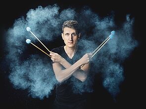 Ein Musiker blickt in die Kamera. In seinen Händen hält er vier Sticks.
