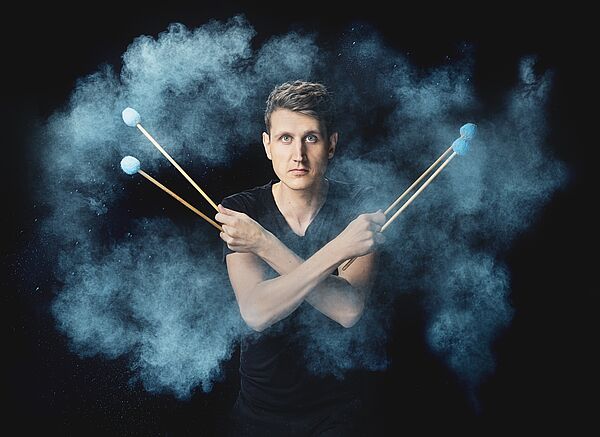 Ein Musiker blickt in die Kamera. In seinen Händen hält er vier Sticks.