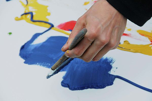 Eine Hand hält einen Pinsel. Auf weißem Untergrund befinden sich blaue, gelbe und rote Farbverläufe.