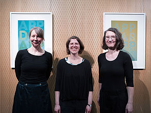 3 Frauen stehen vor einer Wand mit Bildern.