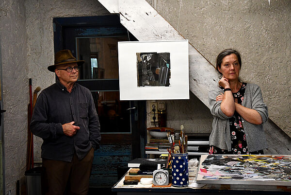 Ein Mann und eine Frau stehen in einem Atelier. Ein Bild hängt zwischen ihnen.