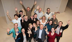 17 Frauen und Männer stehen in einem Raum, strecken die Arme nach oben und blicken lachend in die Kamera.