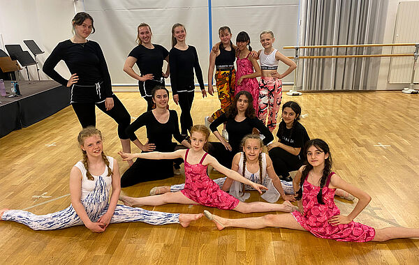 Sieben Kinder und Jugendliche der Tanzgruppe sitzen, zum Teil im Spagat, auf dem Boden. Hinter ihnen stehen sechs Jugendliche.
