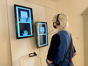 Zwei Bildschirme hängen an einer Wand. Eine Besucherin steht davor und betrachtet die Videos. Sie trägt Kopfhörer.