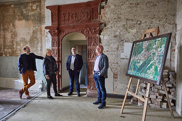 Ein Raum im Schloss. An den Ziegelsteinwänden fehlt Putz. Rechts steht eine Staffelei mit einem Gemälde. Vor einer offenen Tür stehen vier Personen.