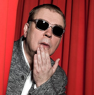 Ein Mann mit einer Sonnenbrille schaut überrascht hinter einem roten Vorhang hervor.