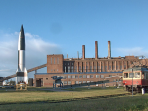 Vor dem Kraftwerk, einem Backsteinbau, stehen ein alter Zug und eine Rakete.