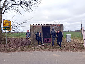 Vier Personen stehen an einer verlassenen Haltestelle aus Steinen. Neben ihn steht ein Ortseingangsschild. Darauf steht: Frauenmark. 