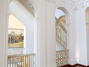 Das Treppenhaus mit weißen Wänden mit Stuck und raumhohen Rundbogenöffnungen. Die Treppengeländer sind weiß und unterschiedlich verschnörkelt.