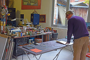 Der Künstler steht an einem Tisch. Vor ihm liegen Zettel. Im Hintergrund steht ein Tisch voller Farb- und Malutensilien.