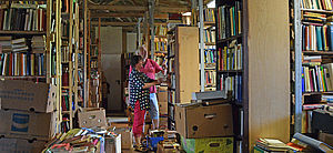 Ein Raum mit vielen Regalen und Kisten. Sie sind gefüllt mit Büchern. Im Raum stehen ein Mann und eine Frau und stöbern in Büchern.
