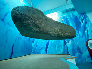 Von der Decke hängt ein Findling herab. Unter dem Findling befindet sich ein Podest, auf das sich Besucher und Besucherinnen legen können. 