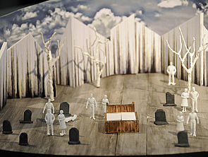 Eine Nahaufnahme vom Bühnenbild. Weiße Wände begrenzen die Kulisse. Auf der Bühne stehen Bäume, Grabsteine, Figuren und ein Doppelbett.