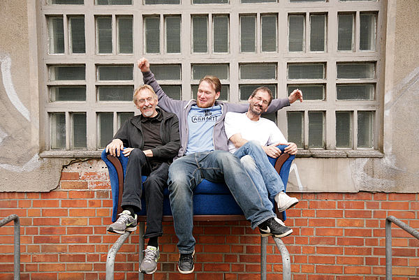 Drei Männer sitzen vor dem Dezernat auf einem blauen Sofa.