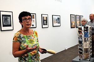 Kornelia Röder steht in einem der Ausstellungsräume. An den Wänden hängen Bilderrahmen. Im Raum steht ein Postkartenständer.