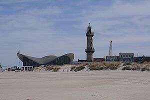 Ein Gebäude mit geschwungenem Dach. Daneben steht ein Leuchtturm. Im Vordergrund liegt der Ostseestrand.