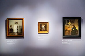 An einer Wand hängen drei Gemälde. 